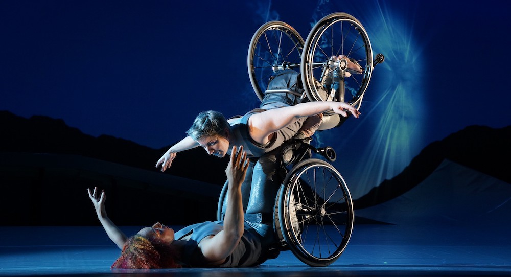 Laurel Lawson as Venus is flying in the air with arms spread wide, wheels spinning, and supported by Alice Sheppard as Andromeda who is lifting from the ground below. They are making eye contact and smiling. Photo by Jay Newman / BRITT Festival.
