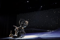 Two dancers in a lift; one balances with arms spread wide, wheelchair wheels spinning, as the other supports her with open arms extended. A starry sky fills the background. Alice Sheppard and Laurel Lawson of Kinetic Light. Photo by BRITT/Jay Newman.