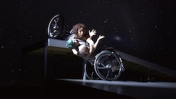 Two dancers, in embrace: one lays on her stomach at the edge of a ramp, holding her partner, as the other leans back. Their wheelchairs shine in the light. Alice Sheppard and Laurel Lawson of Kinetic Light in 'DESCENT'. Photo courtesy of Disability Dance Works/Kinetic Light.