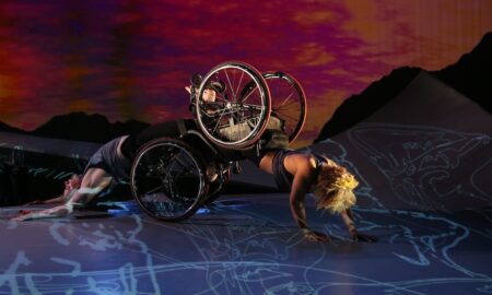 Two dancers, both in wheelchairs; one crawls forward and the other arches her back as she is dragged along the floor. A sunset appears behind them. Alice Sheppard and Laurel Lawson of Kinetic Light. Photo by MANCC/Chris Cameron.