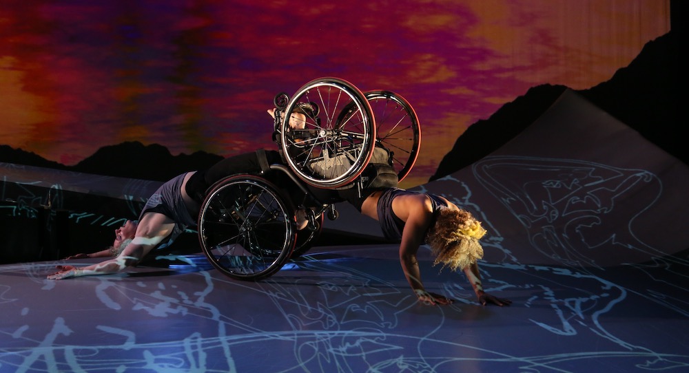 Two dancers, both in wheelchairs; one crawls forward and the other arches her back as she is dragged along the floor. A sunset appears behind them. Alice Sheppard and Laurel Lawson of Kinetic Light. Photo by MANCC/Chris Cameron.