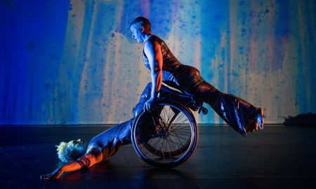 A moment from Kinetic Light’s The Next TiMes. Laurel Lawson holds their body in a diagonal line as they balance atop Alice Sheppard’s wheels and shins. Alice supports from below as they lay back, arms and torso on the ground, hips lifted. Alice is a multiracial Black woman with short curly hair, Laurel is a white person with very short cropped hair; they both wear shimmery sleeveless bodysuits. Behind them, a projection of soft blues, purples, and greens looks like rain on a windowpane. Photo by Cherylynn Tsushima.